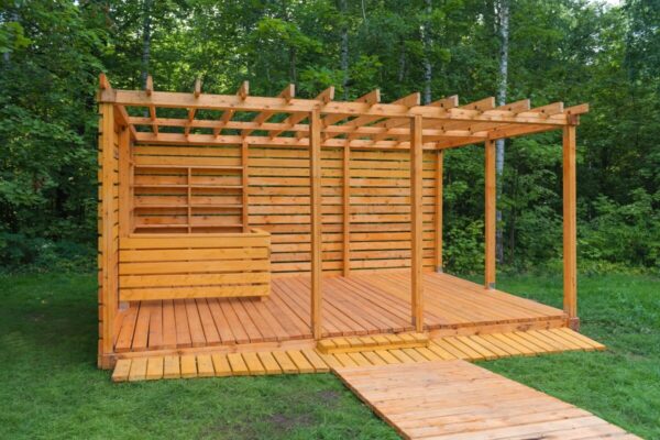 A wooden structure with a pergola and floor made of wooden slats in a forest setting.